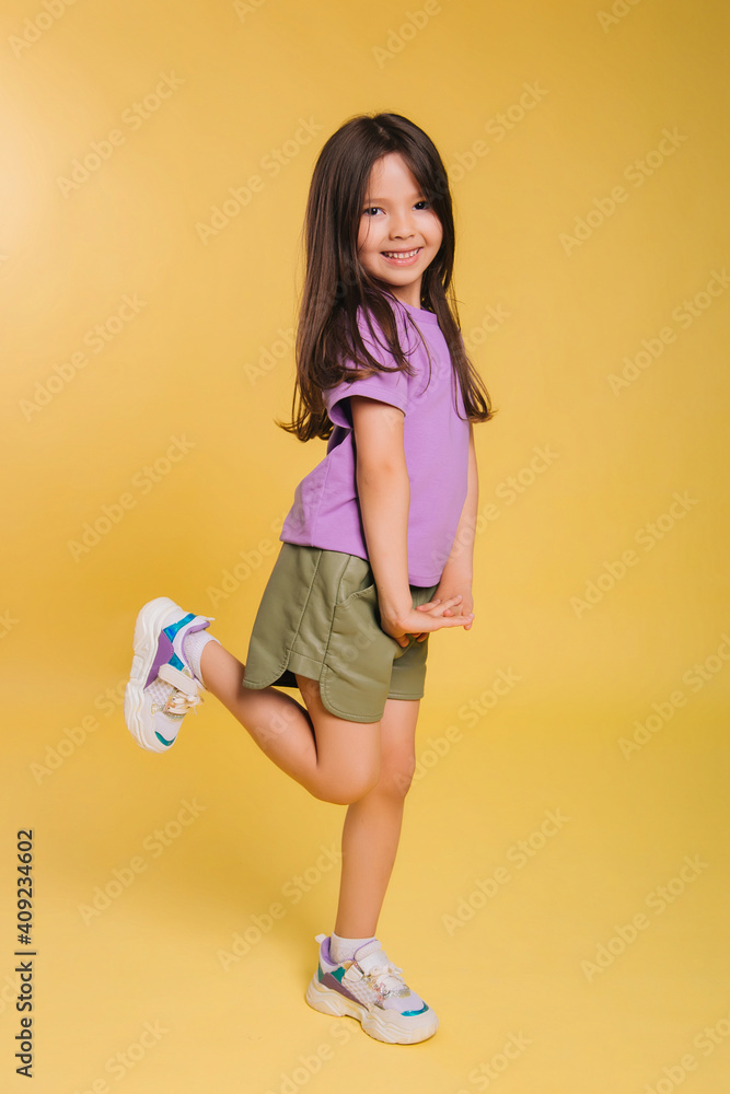 beautiful little girl in a purple t-shirt and shorts posing on a yellow background. A cute baby. Photo in studio beautiful little girl in a purple t-shirt and shorts posing on a yellow background. A cute baby. Photo in studio