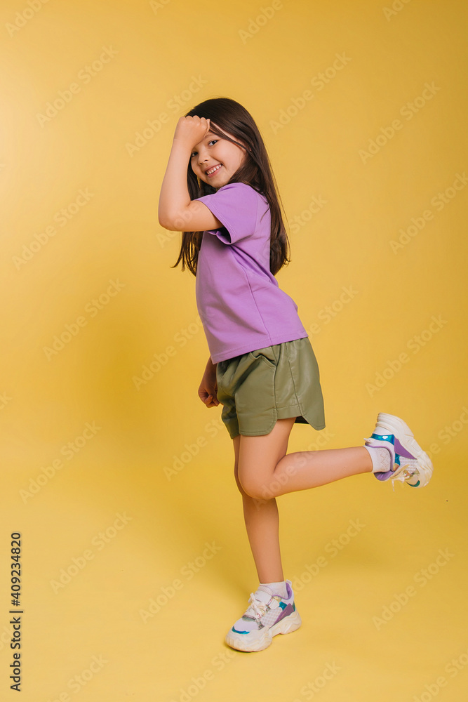 beautiful little girl in a purple t-shirt and shorts posing on a yellow background. A cute baby. Photo in studio beautiful little girl in a purple t-shirt and shorts posing on a yellow background. A cute baby. Photo in studio