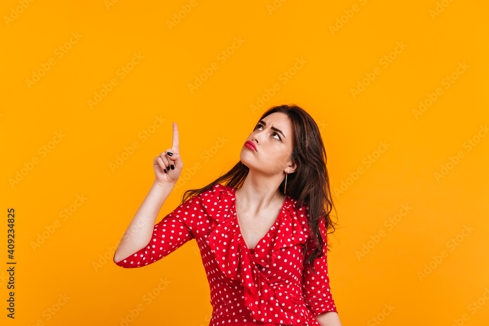 Pensive sad brunette in red top shows finger up. Portrait of girl on isolated yellow background Pensive sad brunette in red top shows finger up. Portrait of girl on isolated yellow background