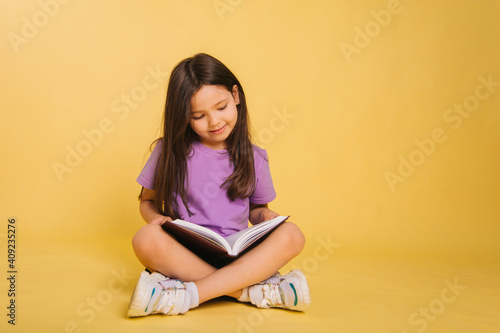 beautiful little girl in a purple t-shirt reads a book while sitting on a yellow background. Cute child learns lessons.