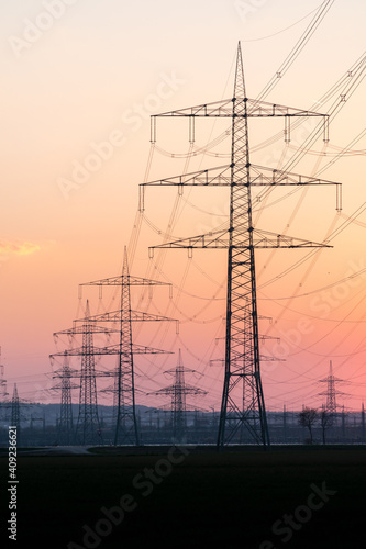 Electricity pylons and electric power transmission lines against vibrant orange sky at sunset. High Voltage towers provide power supply for good infrastructure to rural areas. Vertical format.