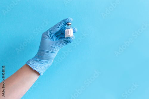 Hand in protective blue gloves hold medicine bottle of vaccine on blue background. Concept of vaccination and healthcare.