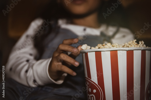 The little girl sat watching a movie in the cinema, eating popcorn in a paper bucket with her delicious hand, and she ate greedily.