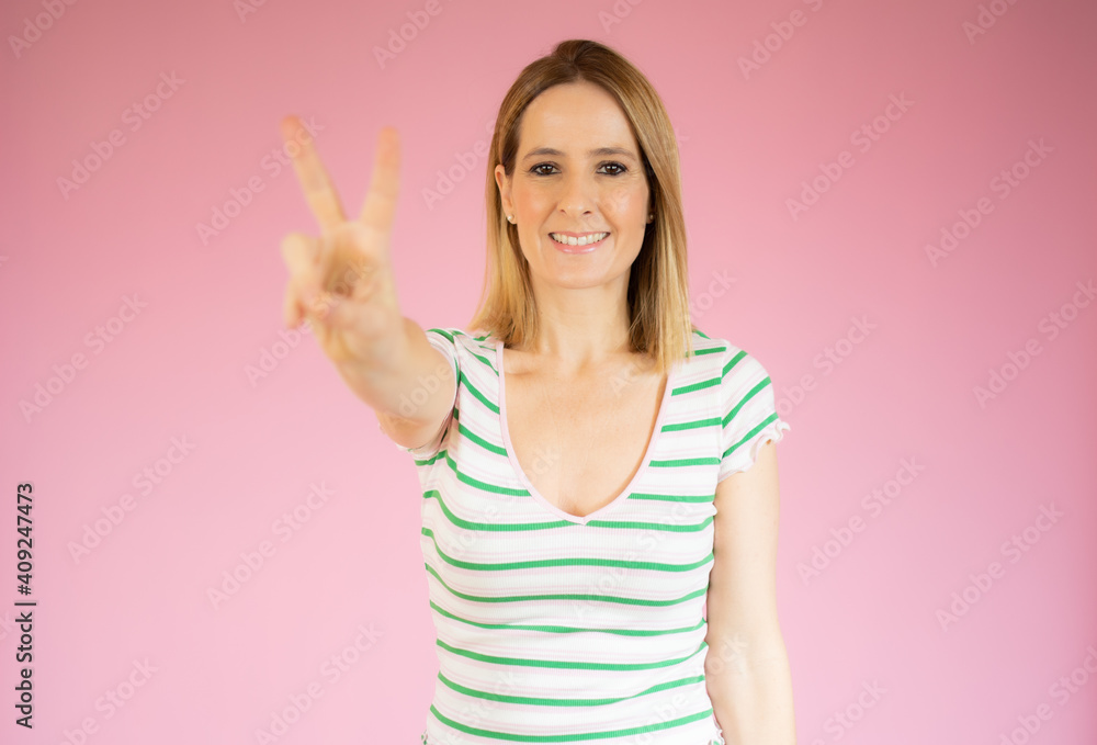 Portrait of a smiling happy woman showing victory sign and looking at camera isolated over pink background Portrait of a smiling happy woman showing victory sign and looking at camera isolated over pink background