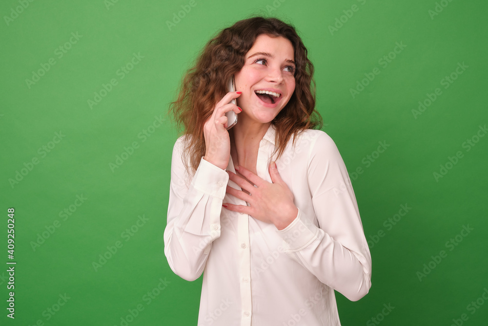 A cheerful girl in white clothes on a green background. The girl holds the phone in her hand and emotionally talks on it, the other hand is pressed to her chest. A cheerful girl in white clothes on a green background. The girl holds the phone in her hand and emotionally talks on it, the other hand is pressed to her chest.