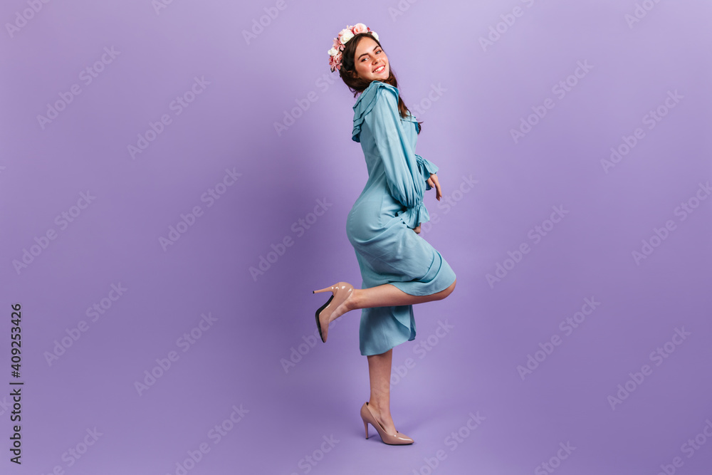 Full-length shot of positive young brunette in heels and midi dress. Female model with flowers in her hair smiling on lilac background Full-length shot of positive young brunette in heels and midi dress. Female model with flowers in her hair smiling on lilac background