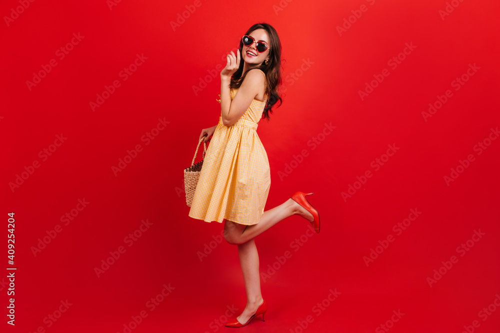 Snapshot full-length of pretty girl in short yellow dress on red background. Woman with dark wavy hair in sunglasses is smiling cute Snapshot full-length of pretty girl in short yellow dress on red background. Woman with dark wavy hair in sunglasses is smiling cute