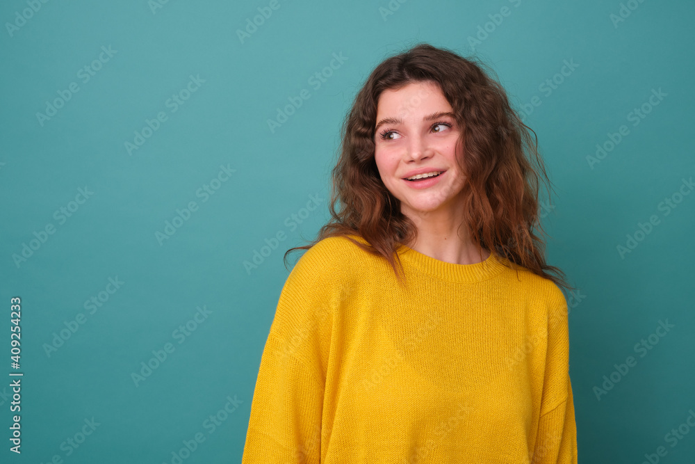 Horizontal shot of a cute young curly girl, has a good mood, feels nice to receive a compliment, focused away, isolated on a blue background Horizontal shot of a cute young curly girl, has a good mood, feels nice to receive a compliment, focused away, isolated on a blue background