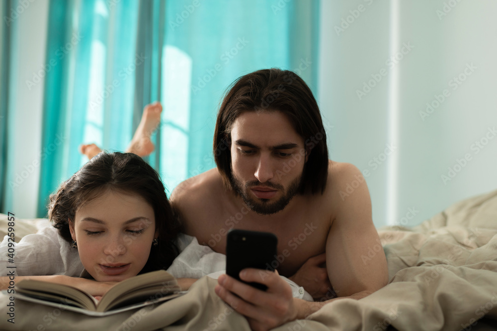 Young Man Looking At Smartphone while his girlfriend Reading Boo Young Man Looking At Smartphone while his girlfriend Reading Boo