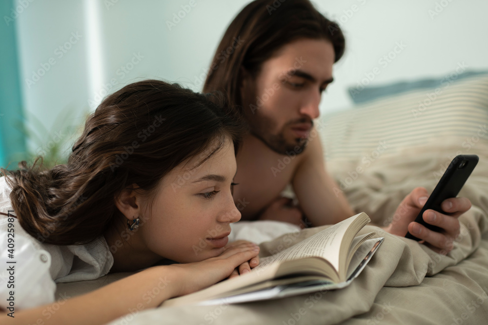 Young Man Looking At Smartphone while his girlfriend Reading Boo Young Man Looking At Smartphone while his girlfriend Reading Boo