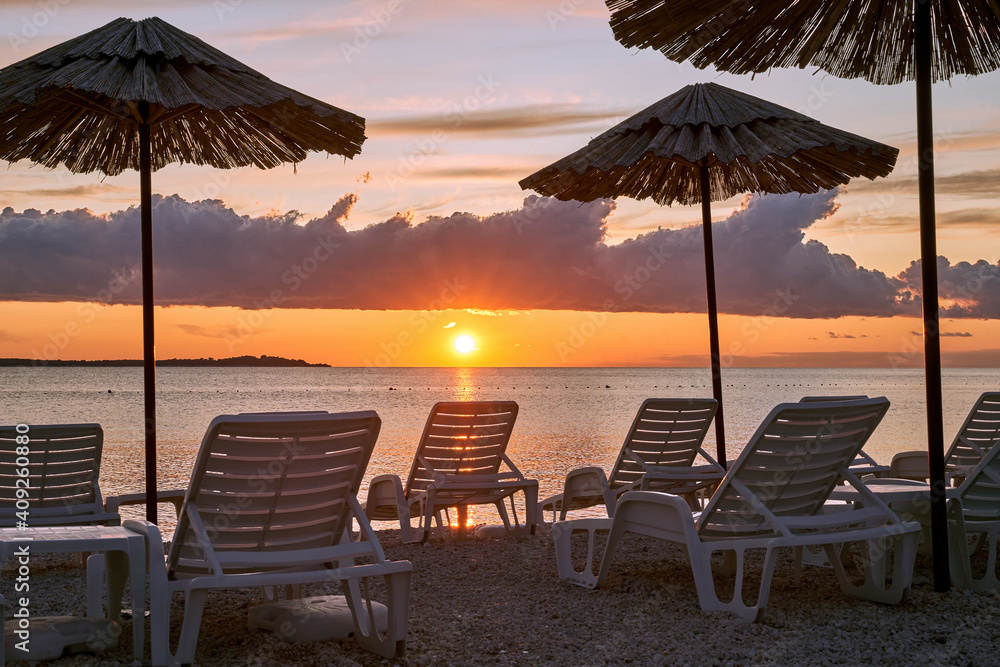 Obraz premium Straw beach umbrellas and chairs on a sunset beach with beautiful clouds