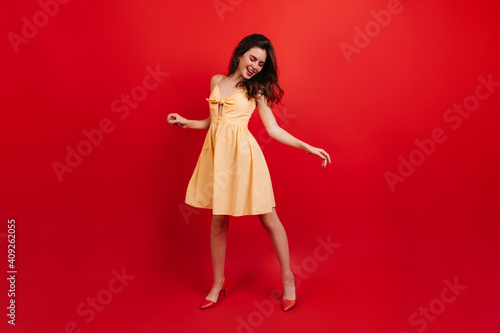 Full-length shot of active woman dancing on red background. Lady in sundress and heels having fun