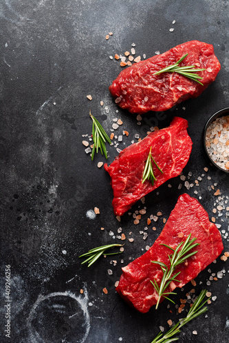 Three Raw beef steak with spices, onions and rosemary on dark slate or concrete background. Top view