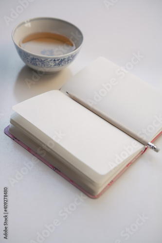 Closeup fo opened notebook with wood pencil on light table, blurred cup of tea in background