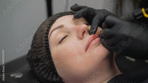 beautiful young girl getting tattoo. Applying permanent makeup to the lips. Beautician applying permanent makeup lipstick to a young woman in a cosmetic center.