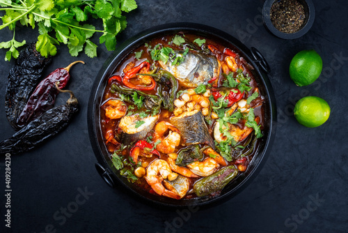 Modern style traditional Mexican seafood pozole soup with fish, king prawns and hominy in a clear sauce served as top view in design pot