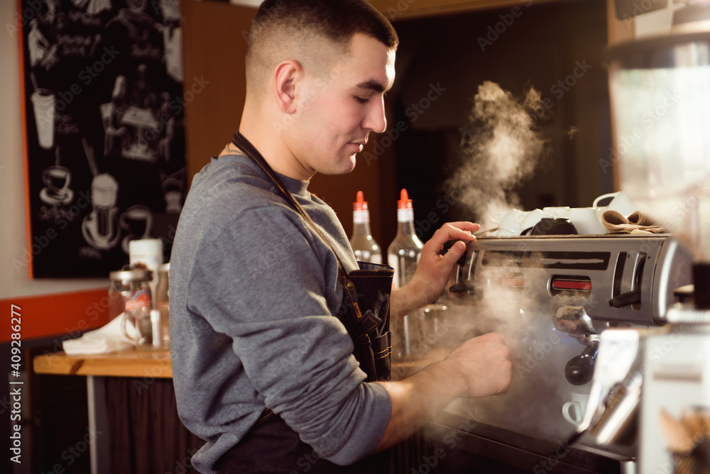 Professional barista holding metal jug warming milk using the coffee machine. Happy young man preparing coffee at counter Professional barista holding metal jug warming milk using the coffee machine. Happy young man preparing coffee at counter