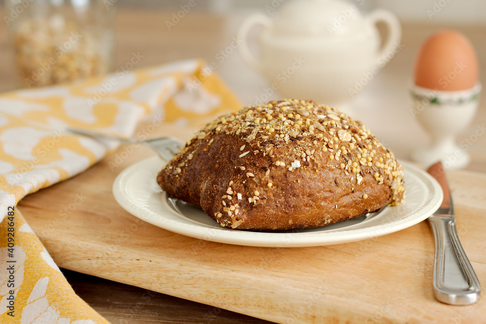 Closeup view of a healthy bun with multigrain mixed cereal seeds on a white porcelain plate with a knife, wooden tray and yellow dishcloth. Tasty breakfast with an boiled egg with selective focus. Closeup view of a healthy bun with multigrain mixed cereal seeds on a white porcelain plate with a knife, wooden tray and yellow dishcloth. Tasty breakfast with an boiled egg with selective focus.