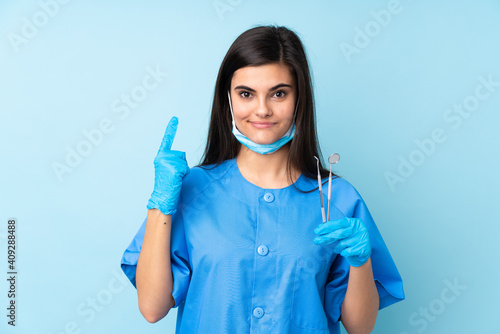 Young woman dentist holding tools over isolated blue background pointing with the index finger a great idea