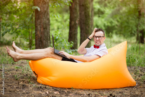 Scared student in glasses with laptop computer lying on inflatable mattress outdoor in summer park. Education, business, fail and technology concept © Svetlana