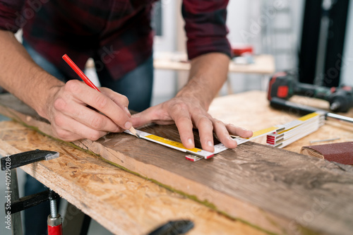 Close-up of a hardworking professional carpenter holding a angular ruler and pencil while measuring a board in a carpentry workshop. A bearded DIY enthusiast measures wood. There are a locksmith table