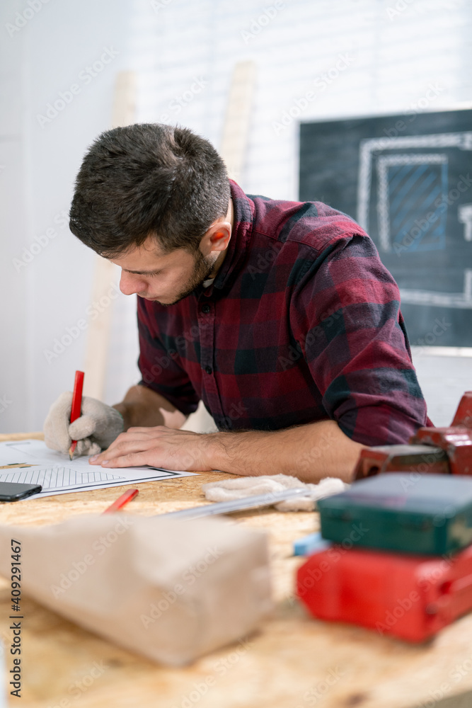 Portrait of a hardworking professional carpenter holding a pencil in a carpentry workshop. A bearded DIY enthusiast doing notes. There are a locksmith table and tools in the background. Portrait of a hardworking professional carpenter holding a pencil in a carpentry workshop. A bearded DIY enthusiast doing notes. There are a locksmith table and tools in the background.