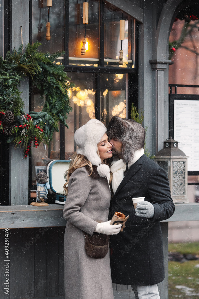 Fashion couple of lovers standing on winter street in snowy weather, man and woman wear fur hat and warm wool coat, hugging, drinking hot drinks. St. Valentines Day, Christmas holidays concept. 