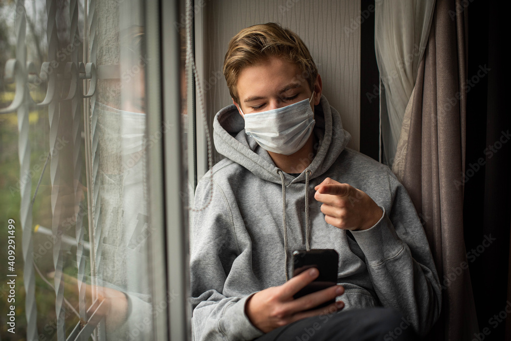 Sad teenage boy with phone confined at home by the coronavirus crisis, looks bored out the window without being able to leave home, defocused background. Sad teenage boy with phone confined at home by the coronavirus crisis, looks bored out the window without being able to leave home, defocused background.