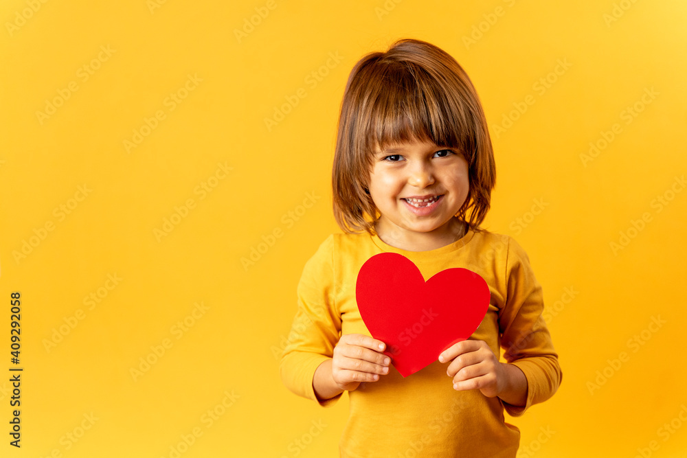 A child with a red heart stands on a yellow background. Little cute boy holds a greeting card in his hand A child with a red heart stands on a yellow background. Little cute boy holds a greeting card in his hand