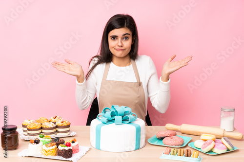 Pastry chef with a big cake in a table over isolated pink background having doubts while raising hands
