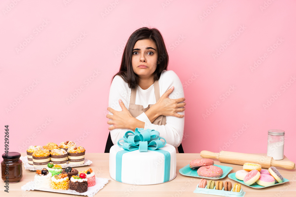 Pastry chef with a big cake in a table over isolated pink background freezing Pastry chef with a big cake in a table over isolated pink background freezing