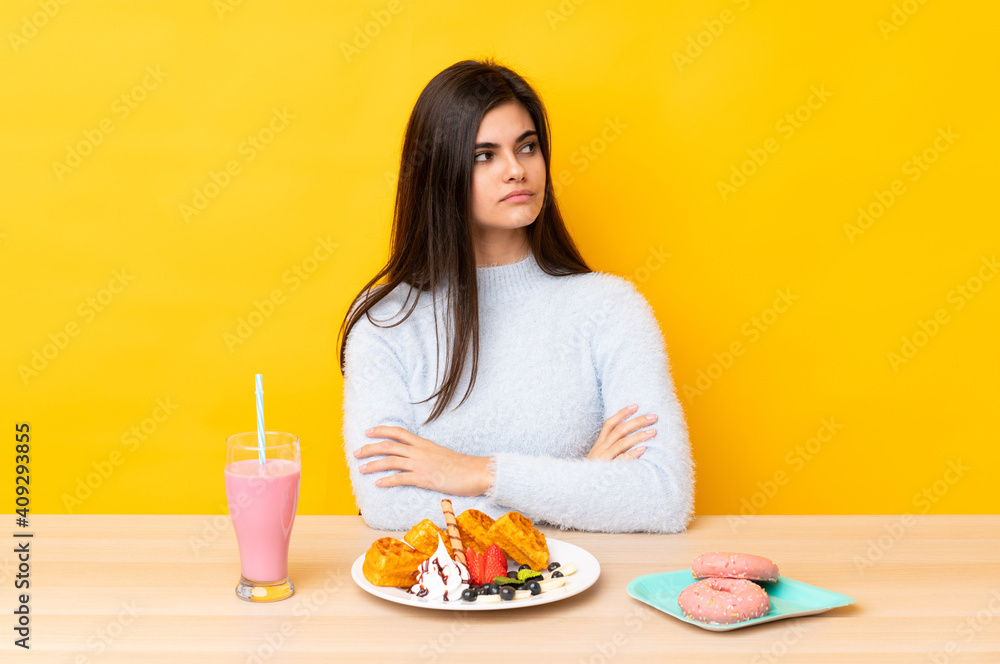 Young woman eating waffles and milkshake in a table over isolated yellow background thinking an idea Young woman eating waffles and milkshake in a table over isolated yellow background thinking an idea