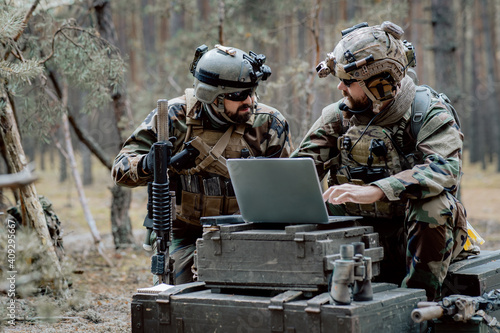 Foto Bearded soldiers in uniform sit on military transport crates, analyze data on a laptop and work out tactics at a temporary forest base