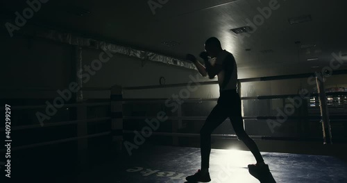 Professional male boxer training in the ring. Professional male boxer training in the ring in a darkened gym throwing punches and practicing his footwork backlit by a bright light
