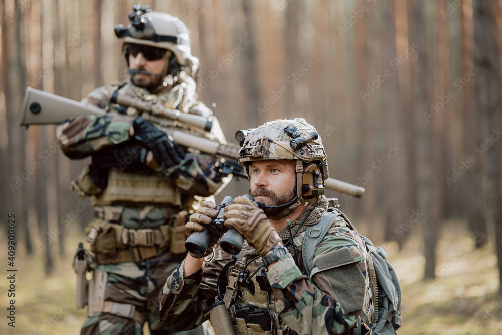 Portrait of a bearded middle-aged soldier in a Woodland military uniform and a helmet with headphones on his head, holding a rifle and looking around through the thick pine woods.