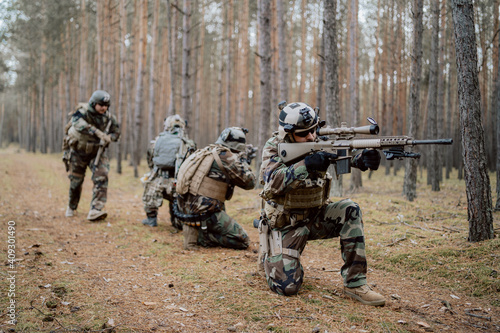 Squad of Four Fully Equipped Soldiers in Camouflage on a Reconnaissance Military Mission, Aiming Rifles. They're Moving in Formation Through Dense Pine Forest. photo