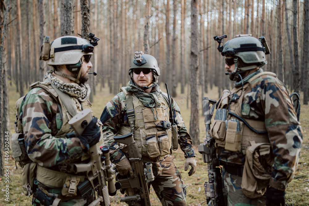 A group of middle-aged bearded soldiers in uniforms and tactical vests discusses the action plan and prepares for action at a temporary forest base. The commander using military tablet.