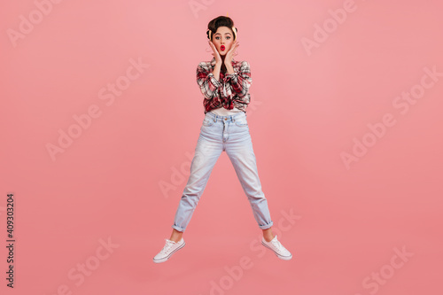 Amazed woman in jeans jumping on pink background. Pretty pinup girl in checkered shirt expressing surprised emotions.