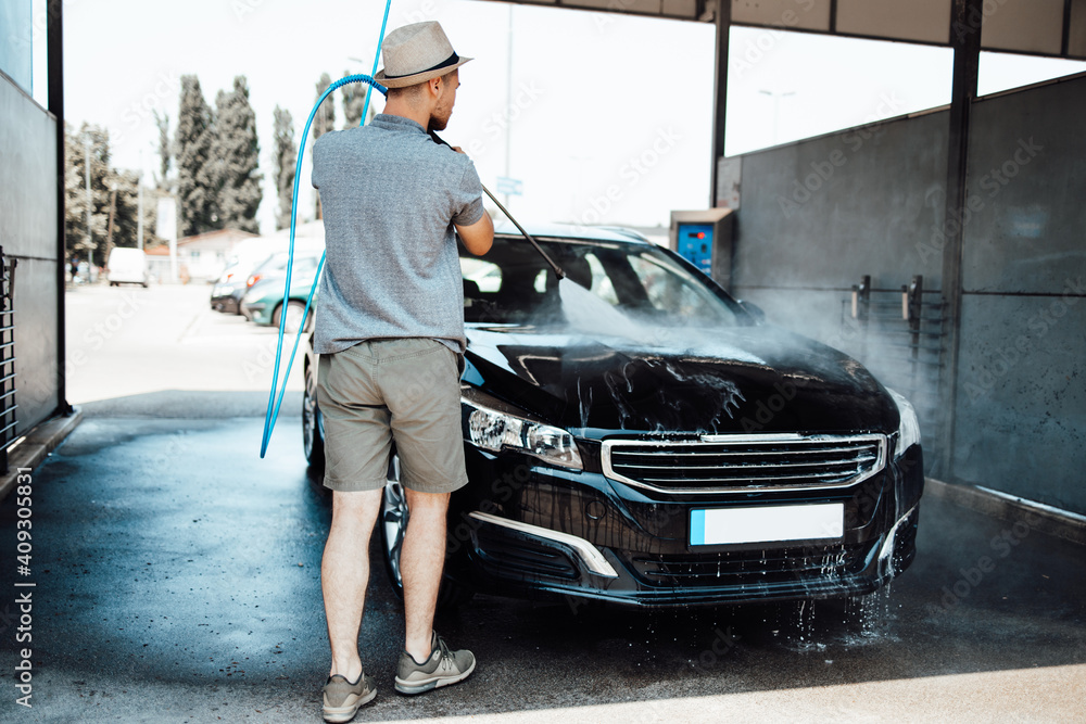 Fototapeta premium Young man with hat washing his car during daylight at car wash station using high pressure water.