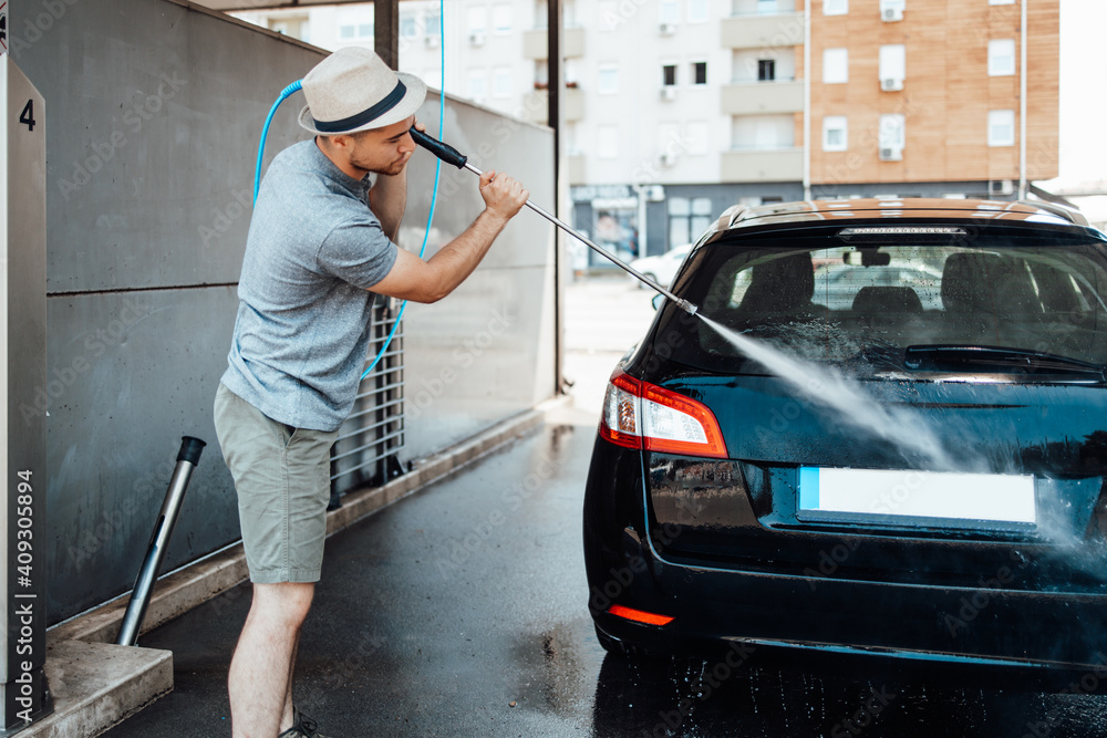 Young man with hat washing his car during daylight at car wash station using high pressure water.