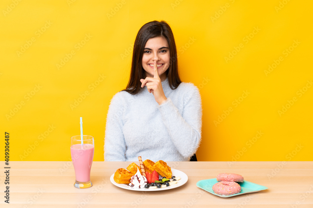 Young woman eating waffles and milkshake in a table over isolated yellow background doing silence gesture Young woman eating waffles and milkshake in a table over isolated yellow background doing silence gesture
