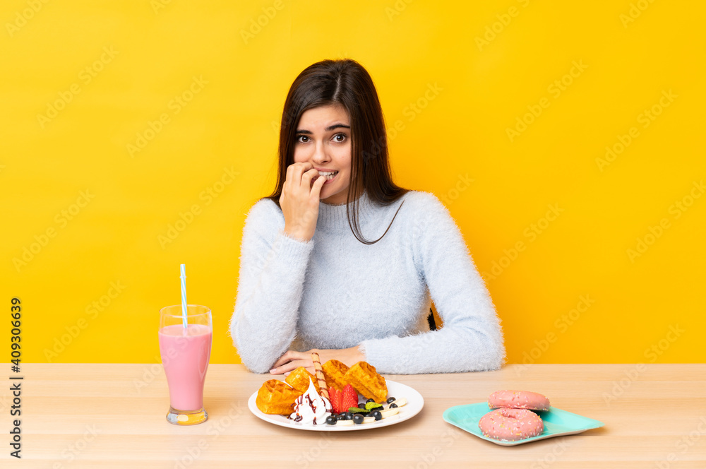 Young woman eating waffles and milkshake in a table over isolated yellow background nervous and scared Young woman eating waffles and milkshake in a table over isolated yellow background nervous and scared