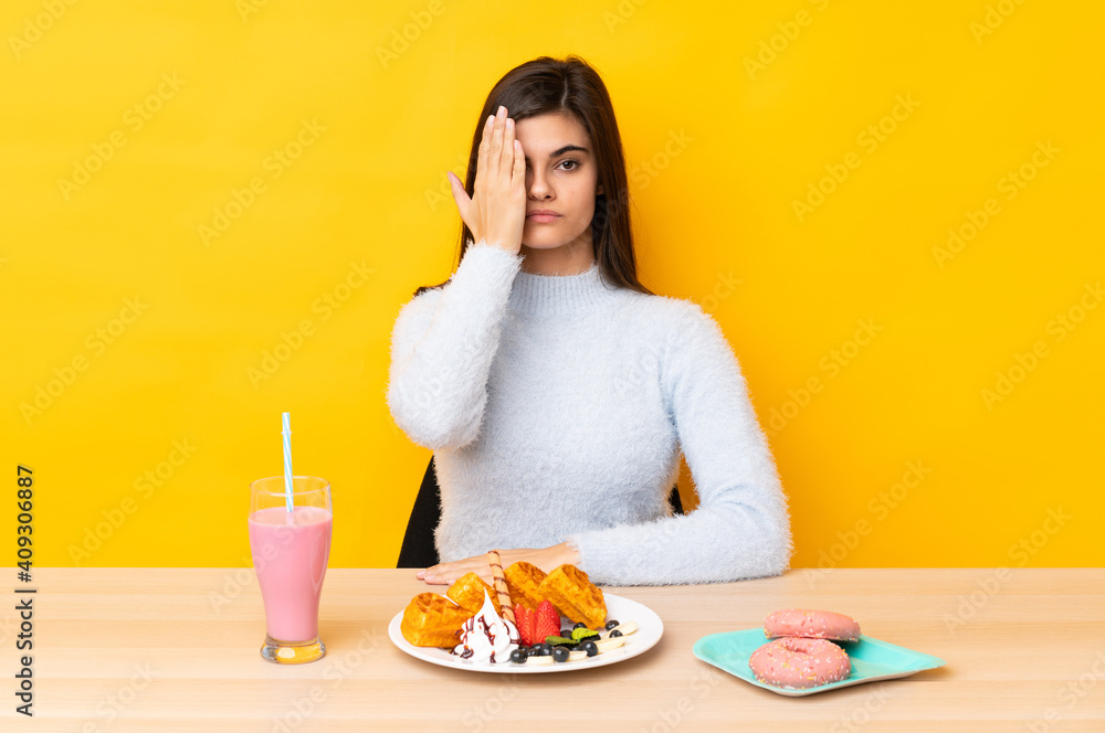 Young woman eating waffles and milkshake in a table over isolated yellow background covering a eye by hand