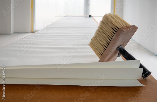 Wide paint brush for glue with wooden base for bristle,black handle,sheet of white wallpaper unfolded in length on table for pasting against renovating room with french window in backgdrop.Copy space. © Nina