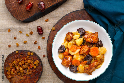 A dish of Jewish cuisine sweet tsimes with carrots, dates and turkey meat in a white plate on a round board on a linen background near dried fruits. photo