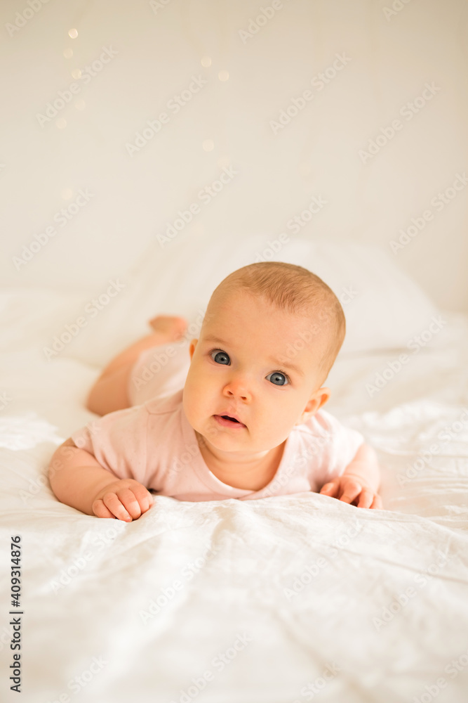 a little baby girl lies in a pink bodysuit on a white blanket on the bed and looks at the camera