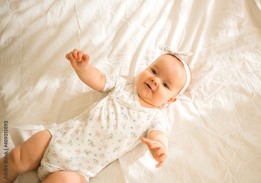 joyful little baby girl lies in a colored bodysuit and a headband on a white blanket on the bed and looks at the camera joyful little baby girl lies in a colored bodysuit and a headband on a white blanket on the bed and looks at the camera