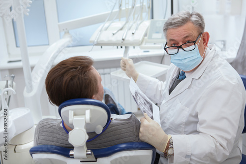 Senior male dentist wearing medical face mask, looking to the camera during dental appointment with patient