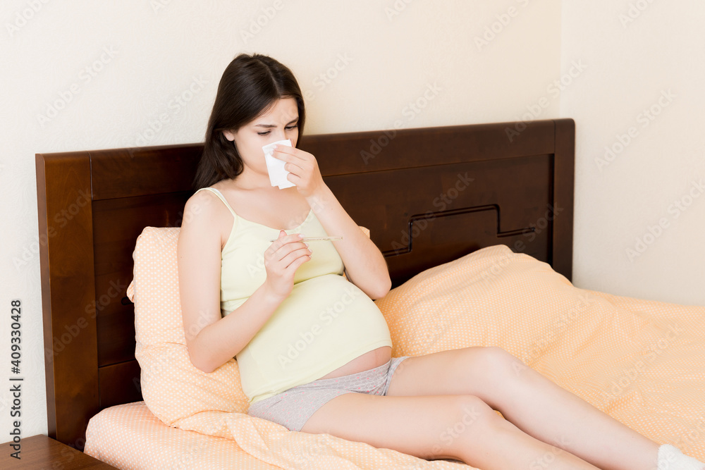 Portrait of young unhappy pregnant woman leaning on pillows on bed and blowing her nose into tissue. Future mom caught cold and sneezing Portrait of young unhappy pregnant woman leaning on pillows on bed and blowing her nose into tissue. Future mom caught cold and sneezing