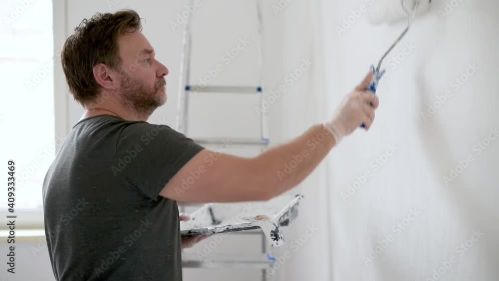 Portrait of a handsome mature man making repairs in the apartment. The person paints the wall white with roller. Do it yourself. DIY. Repair with their hands. Portrait of a handsome mature man making repairs in the apartment. The person paints the wall white with roller. Do it yourself. DIY. Repair with their hands.